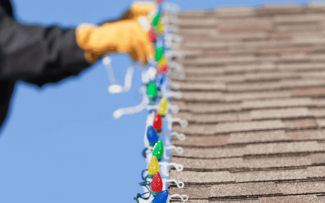 hands wearing gloves and a black long sleeved shirt hanging holiday lights on the eave of a home showing the lights clipped to the edge and brown shingles